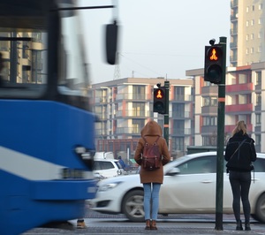 W centralnej części kadru widać dwie osoby piesze czekające przed przejściem dla pieszych na zmianę sygnału czerwonego zakazującego wejścia. W tle przejeżdżające po jezdni pojazdy. Z lewej strony kadru widać przejeżdżający tramwaj. Pogoda słoneczna, ubiór pieszych wskazuje na czas zimowy.