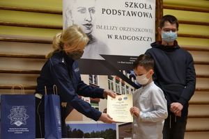 Policjantka wręcza dyplom jednemu uczniowi, drugi chłopiec stoi obok. Wszyscy ustawieni są na tle szkolnego baneru.