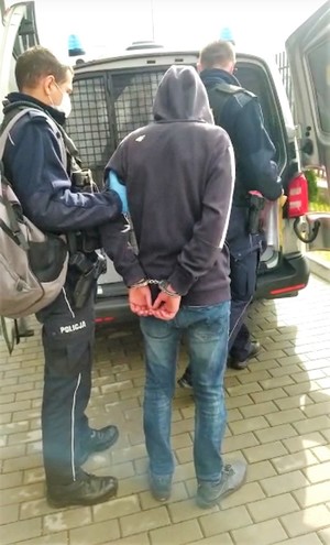 Dwóch umundurowanych policjantów którzy prowadzą zatrzymanego mężczyznę który na rekach trzymanych z tyłu ma założone kajdanki_