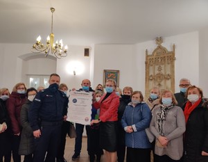 policjant z seniorami stojący twarzą do zdjęcia, policjant z jednym z seniorów trzymają wspólnie  duża ulotkę informacyjna o bezpieczeństwie seniorów