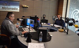 Redaktor radia przeprowadza wywiad z policjantem w studiu radiowym. W tle wiszące na ścianie logo radia „ GO+ Motoradio”. Widoczne mikrofony i urządzenia techniczne studia.