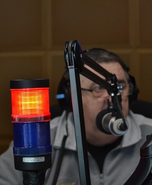 Na pierwszym planie – Zapalona czerwona lampka sygnalizująca wejście na antenę. Na drugim planie mikrofon oraz redaktor prowadzący wywiad.
