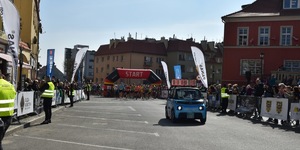 Na rynku miasta odbywa się start zawodników do półmaratonu. Z przodu przed zawodnikami samochód pilotujący bieg koloru niebieskiego.