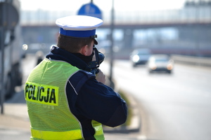Policjant ruchu drogowego  - widoczna biała czapka, żółta kamizelka odblaskowa z napisem POLICJA -  dokonujący pomiaru prędkości. W tle widać nadjeżdżające pojazdy