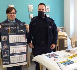 Umundurowany policjant stojący obok kobiety prezentującej kalendarz na 2022 roku z informacją jak nie dać oszukać.