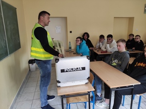 technik kryminalistyki w odblaskowej kamizelce rozmawia z uczniami o swojej pracy. Uczniowiie siedzą w szkolnych ławkach.