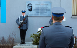 Policjant kompanii honorowej niesie wieniec pod tablice Ludwika Drożańskiego