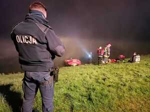 policjant stojący tyłem na brzegu, w tle strażacy wyciągają z wody pojazd