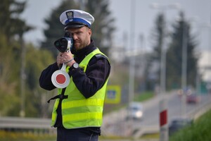 Policjant ruchu drogowego (biała czapka, żółta kamizelka), stojąc przy drodze mierzy prędkość pojazdów za pomocą ręcznego miernika. W lewej dłoni trzyma też tarczę do zatrzymywania pojazdów).
