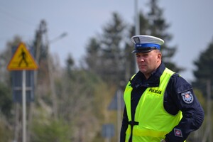 Policjant ruchu drogowego (biała czapka, żółta kamizelka) nadzoruje ruch na drodze. W tle widać znak ostrzegawczy „uwaga przejście dla pieszych”