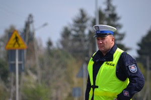 Policjant ruchu drogowego (biała czapka, żółta kamizelka) nadzoruje ruch na drodze. W tle widać znak ostrzegawczy „uwaga przejście dla pieszych”