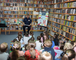 Na pierwszym tle dzieci. W dalszym tle policjant, półki z książkami oraz tablica
