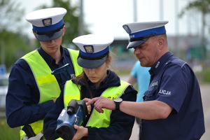 Policjant prowadzący szkolenie objaśnia policjantce obsługę ręcznego miernika prędkości. Czynności przygląda się również drugi policjant.