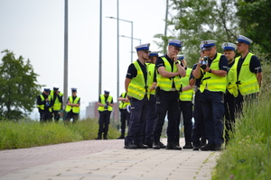 Dwie grupy policjantów ruchu drogowego (białe czapki, żółte kamizelki odblaskowe), stojących na chodniku drogi, zapoznających się z obsługą ręcznych mierników prędkości.