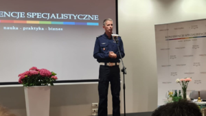 policjant przed mikrofonem stoi na tle ekranu wyświetlającego tytuł konferencje specjalistyczne