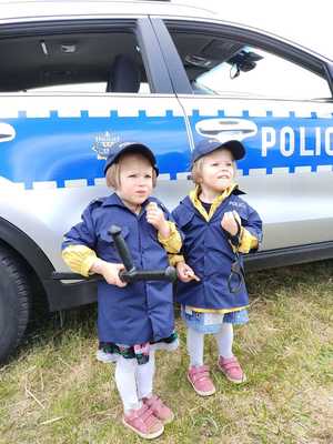 Dwie małe dziewczynki ubrane w strój policjanta stojące na tle rdiowozu.