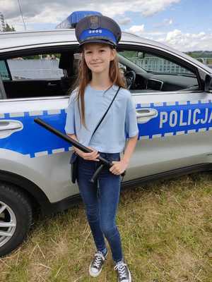 Dziewczynka z policyjną czapką na głowie. W rękach trzyma policyjną pałkę. Stoi na tle radiowozu.