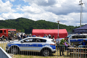 Miejsce imprezy plenerowej. W tle namioty, oznakone radiowozy, wóz strażacki oraz tłum osób.