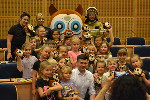 Grupa dzieci wraz z Panem Marszałkiem Smółką, policjantką, panią sekretarz, strażakiem oraz maskotką „Sówką”