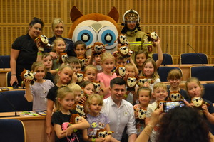 Grupa dzieci wraz z Panem Marszałkiem Smółką, policjantką, panią sekretarz, strażakiem oraz maskotką „Sówką”