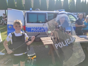 Dwóch chłopców stoi ubranych w policyjny strój ochronny, jeden jest w kasku i trzyma w ręku tarczę, a drugi pałkę. Za nimi radiowóz policyjny i policjant rozmawiający z kobietą.