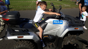 Dwóch policjantów stoi przy radiowozie i rozmawiają z kobietą, obok na policyjnym quadzie siedzi młody chłopiec