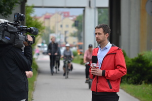 Dziennikarz TVP INFO stoi przed nagrywającym go kamerzystą. W tle – osoby jadące na rowerach i hulajnodze.