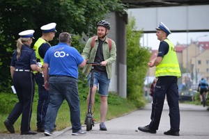 Osoba kierująca hulajnogą stoi przy krawędzi drogi dla rowerów i udziela wywiadu dziennikarzowi z GO+ Motoradio. Obok stoją policjanci ruchu drogowego.