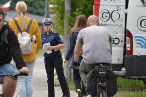 Policjantka ruchu drogowego (widoczna biała czapka) stoi przy drodze dla rowerów i wręcza przejeżdżającym użytkownikom hulajnóg specjalnie przygotowane ulotki z przepisami. Widoczne duże natężenie ruchu.