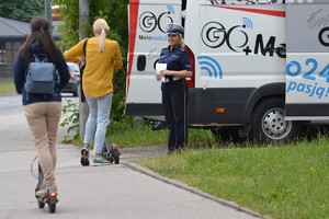 Policjantka ruchu drogowego rozmawia z kobietą kierującą hulajnogą elektryczną. Obok inna kobieta przejeżdża koło nich również hulajnogą elektryczną.