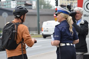 Policjantka ruchu drogowego rozmawia z mężczyzną kierującym hulajnogą elektryczną. W tle widać kamerzystę nagrywającego sytuację.