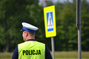 Policjant ruchu drogowego stoi przy przejściu dla pieszych ( w tle znak drogowy - przejście dla pieszych) i nadzoruje ruch