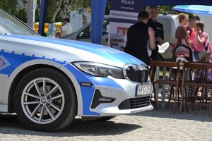 Przedni prawy narożnik radiowozu oznakowanego BMW. W tle stoisko profilaktyczne WRD KWP w Krakowie z policjantką odpowiadającą na pytania