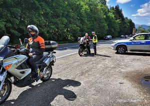 motocyklista siedzi na mototocyklu na poboczu drogi, w tle policjant z drogówki kontroluje kolejny motocykl.na poboczu stoi zaparkowany radiwoóz