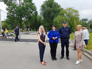 policjant, przedstawicielka starostwa powiatowego i dwie kobiety, stojący przodem do zdjęcia, za nimi w tle miasteczko rowerowe