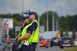 Dwóch policjantów ruchu drogowego (białe czapki, żółte kamizelki) stojąc przy drodze nadzoruje ruch. W tle duże natężenie ruchu, słoneczna, letnia pogoda.