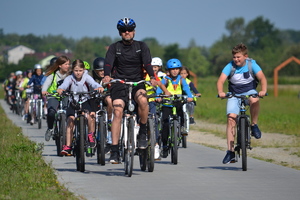 Peleton rowerzystów z dziećmi jadącymi po ścieżce rowerowej, na czele jedzie opiekun. Słoneczna, wakacyjna aura.