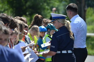 Policjantka ruchu drogowego wraz z Marszałkiem wręczają książeczki edukacyjne dzieciom przybyłym na prelekcjęw plenerze.