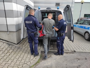 Zatrzymany mężczyzna doprowadzany do radiowozu przez dwóch umundurowanych policjantów. W tle widać budynek komendy i inny radiowóz.
