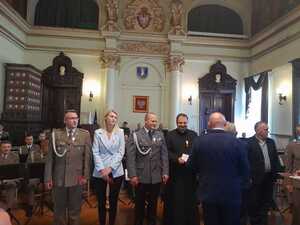 Odznaczeni honorową odznaką policjanci z Brzeska obok nich stojący przedstawiciele wojska i osoby cywilne: kobieta, mężczyzna i ksiądz.