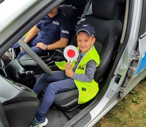 chłopiec w kamizelce odblaskowej, policyjnej czapce i z tarczą do zatrzymywania pojazdów siedzi we wnętrzu radiowozu