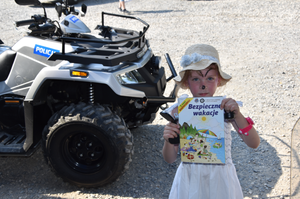Quad z napisem POLICJA. Przed pojazdem dziecko trzymające w rękach książkę reklamującą akcję „Bezpieczne wakacje”.