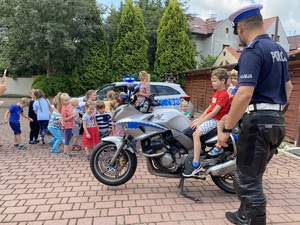 policjant ruchu drogowego przy motocyklu, na którym siedzi dziecko, wokół dzieci uczestniczące w spotkaniu profilaktycznym
