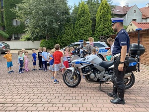 policjant ruchu drogowego przy policyjnym motorze, policyjny radiowóz i dzieci biorące udział w spotkaniu profilaktycznym
