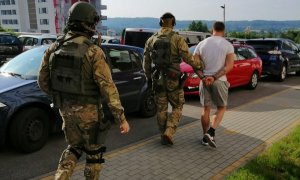 funkcjonariusze z Samodzielnego Pododdziału Kontterrorystycznego Policji KWP w Rzeszowie prowadzą zatrzymanego ubranego w krótkie spodnie i białą koszulkę.  W tle znajdują się pojazdy osobowe