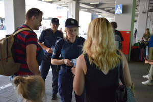 policjantka wraz z policjantem na terenie dworca autobusowego wręczają kobiecie ulotkę informacyjną. obok stoi mężczyzna z dzieckiem