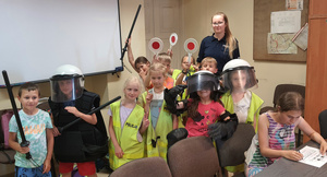 w jednym z pokoi dzieci w kamizelkach odblaskowych trzymają tarcze do zatrzymywania pojazdów, za dziećmi stoi policjantka. Wszyscy pozują do zdjęcia.jpg