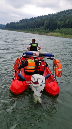 Na wodzie łódź strażacka , na któej siedzą tyłem strażacy oraz policjant w czarnej kamizelce z napisem Policja