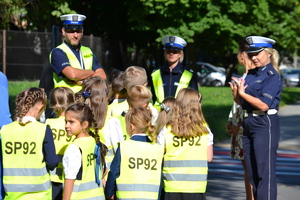 Dzieci w kamizelkach odblaskowych z napisem SP92 słuchają prelekcji prowadzonej przez policjantów ruchu drogowego przy przejściu dla pieszych