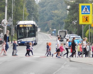 Grupa dzieci idąca w prarach pod opieką kilku osób dorosłych przechodzi przez oznakowane tzw. agatką przejście dla pieszych. W tle nadjeżdżający autobus MPK
